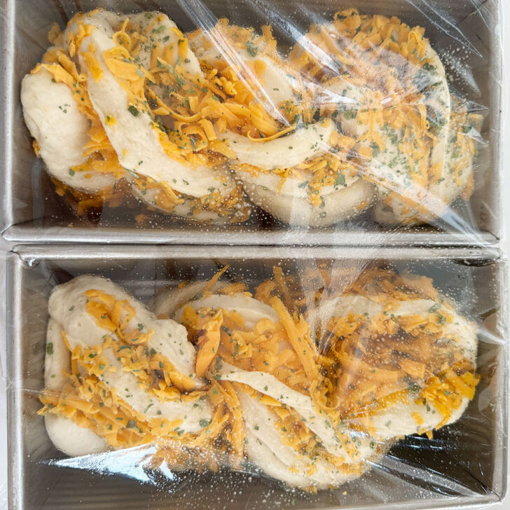 Both cheese bread loaves in pans, covered with plastic wrap.