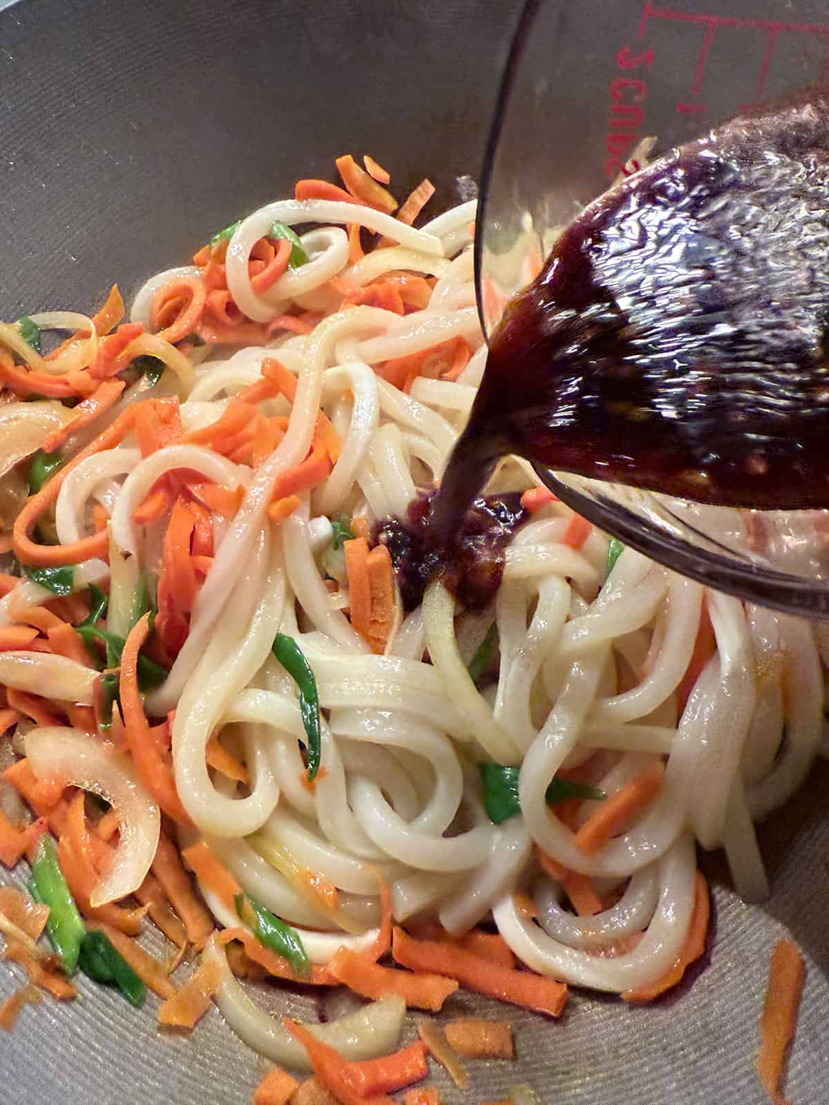 Adding the spicy stir fry sauce to the wok with the udon noodles and vegetables.
