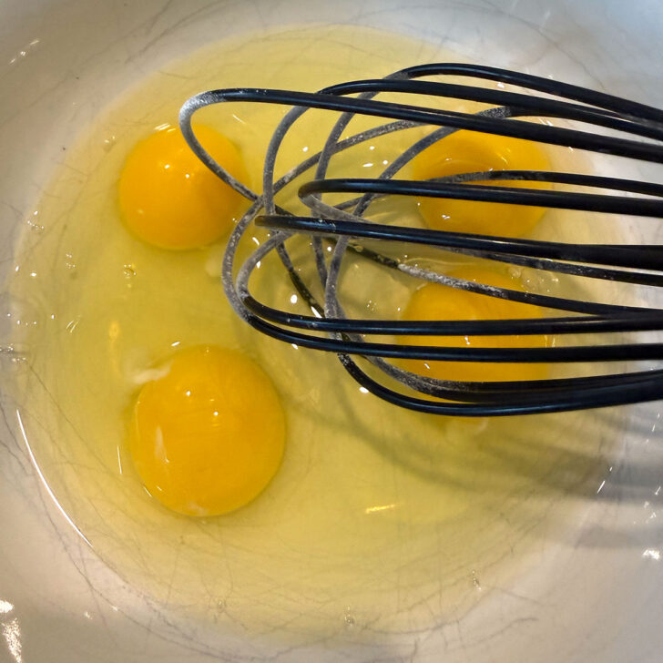 Four eggs in a mixing bowl with a whisk.