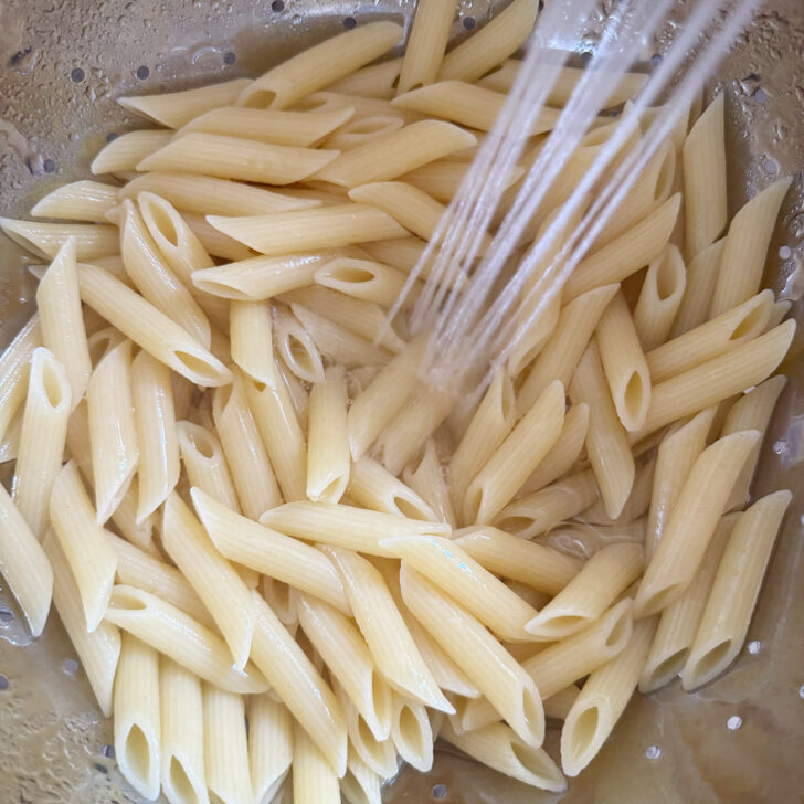 Rinsing cooked penne pasta in a colander after cooking.
