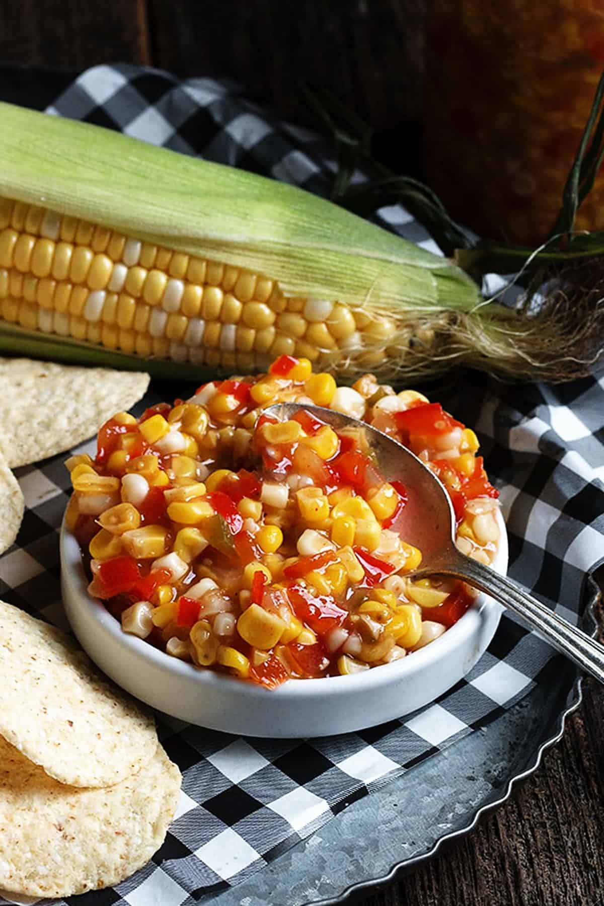 corn and chile salsa in small bowl with tortilla chips and corn in the background