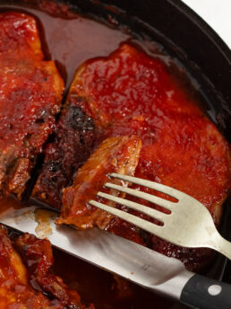 Oven BBQ pork chops in a cast iron skillet with a knife and fork.