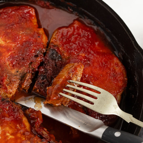 Oven BBQ pork chops in a cast iron skillet with a knife and fork.