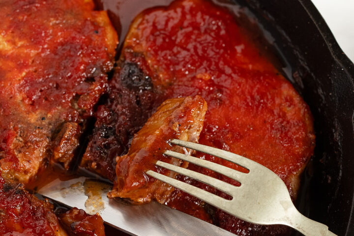 Oven BBQ pork chops in a cast iron skillet with a knife and fork.