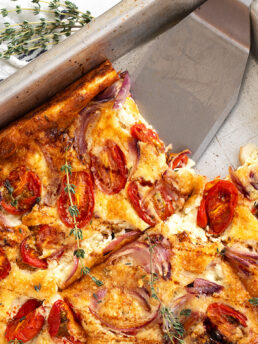 A baked tomato Dutch baby in a metal pan, topped with roasted cherry tomatoes, red onions, and herbs. A slice has been cut and lifted with a metal spatula, while fresh thyme serves as garnish.