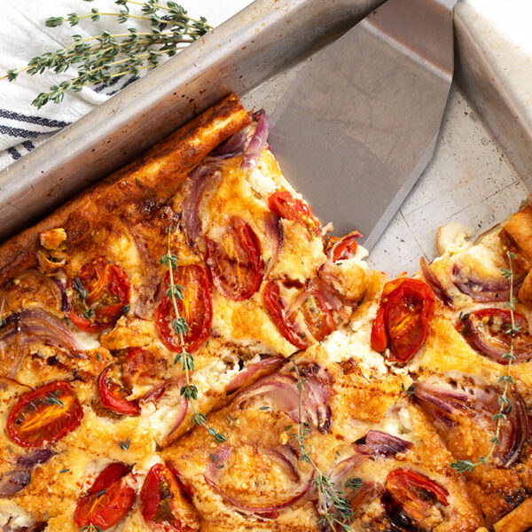 A baked tomato Dutch baby in a metal pan, topped with roasted cherry tomatoes, red onions, and herbs. A slice has been cut and lifted with a metal spatula, while fresh thyme serves as garnish.