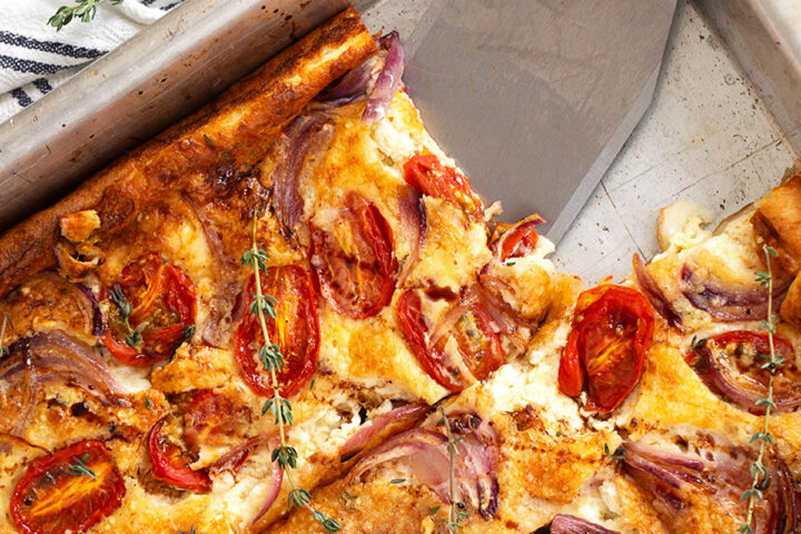 A baked tomato Dutch baby in a metal pan, topped with roasted cherry tomatoes, red onions, and herbs. A slice has been cut and lifted with a metal spatula, while fresh thyme serves as garnish.