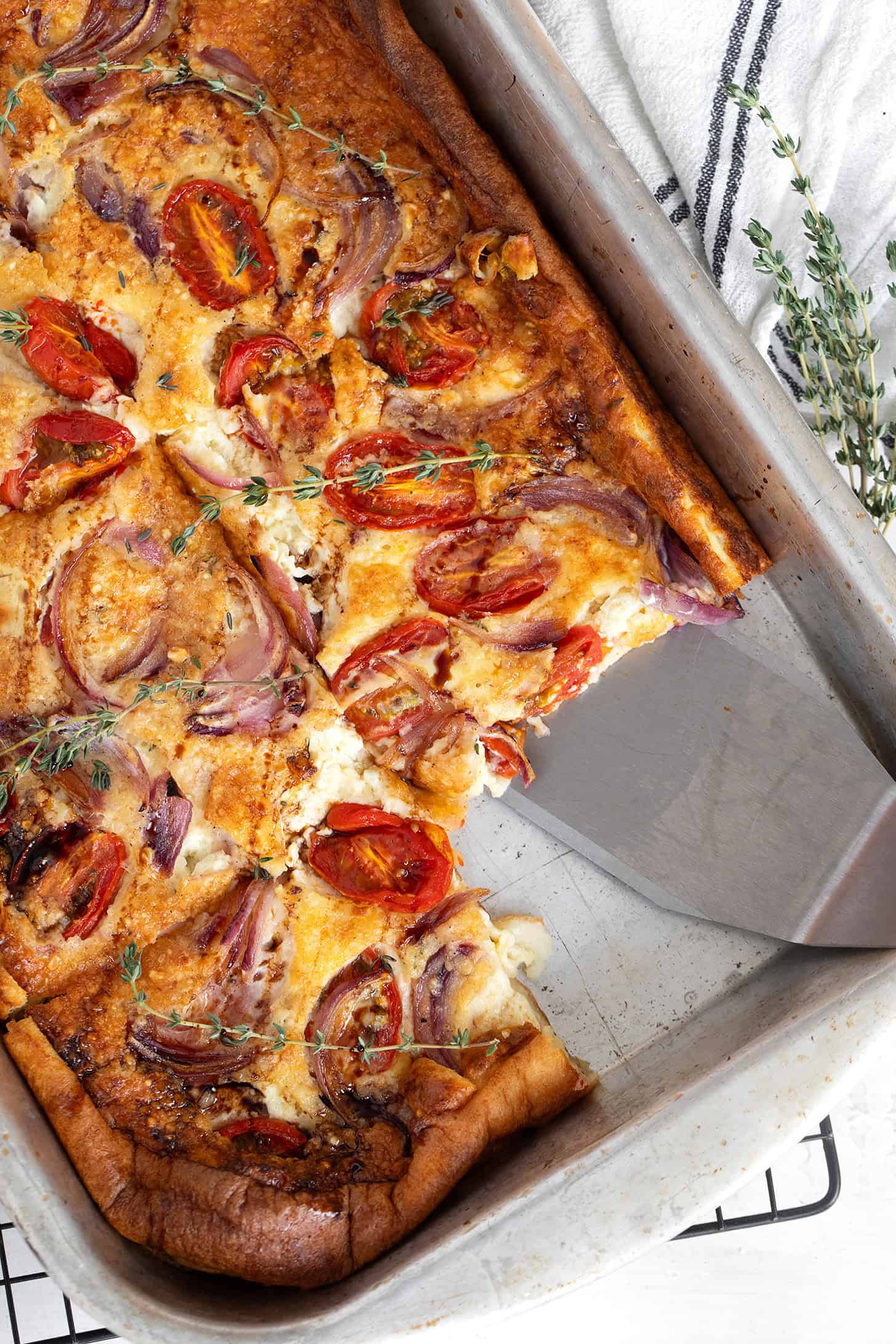 A baked tomato Dutch baby or frittata y tomatoes, red onions, and herbs. A slice has been cut and lifted with a metal spatula, while fresh thyme serves as garnish.