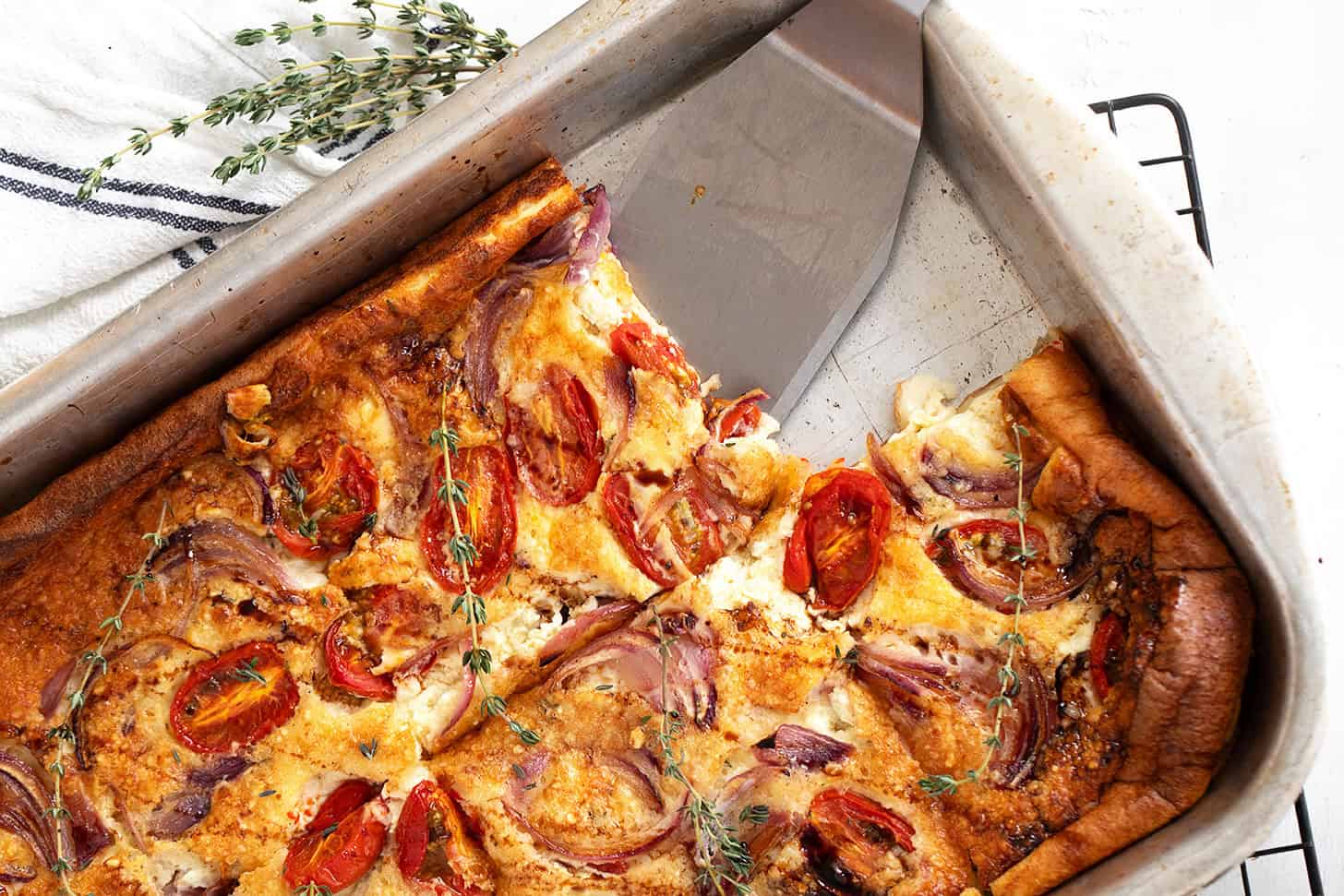 A baked tomato Dutch baby in a metal pan, topped with roasted cherry tomatoes, red onions, and herbs. A slice has been cut and lifted with a metal spatula, while fresh thyme serves as garnish.