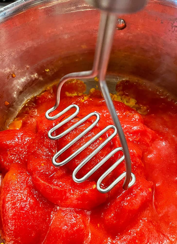 mashing whole canned tomatoes with a potato masher