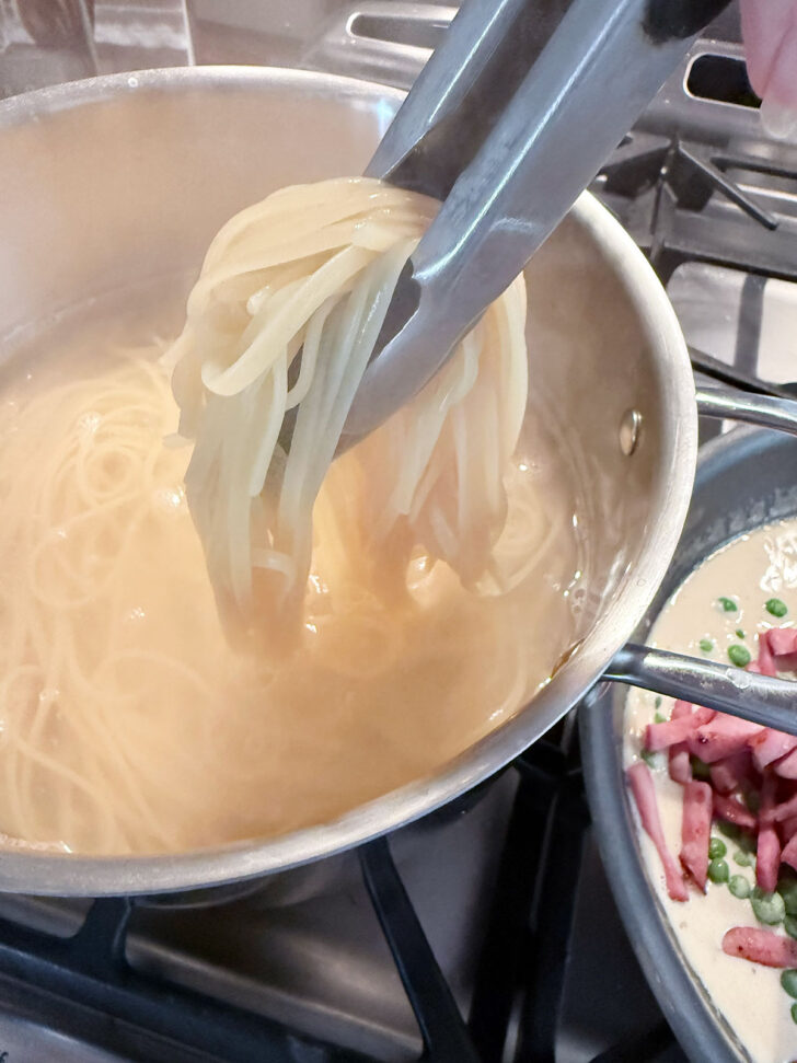 Using tongs to transfer the cooked pasta to the skillet.