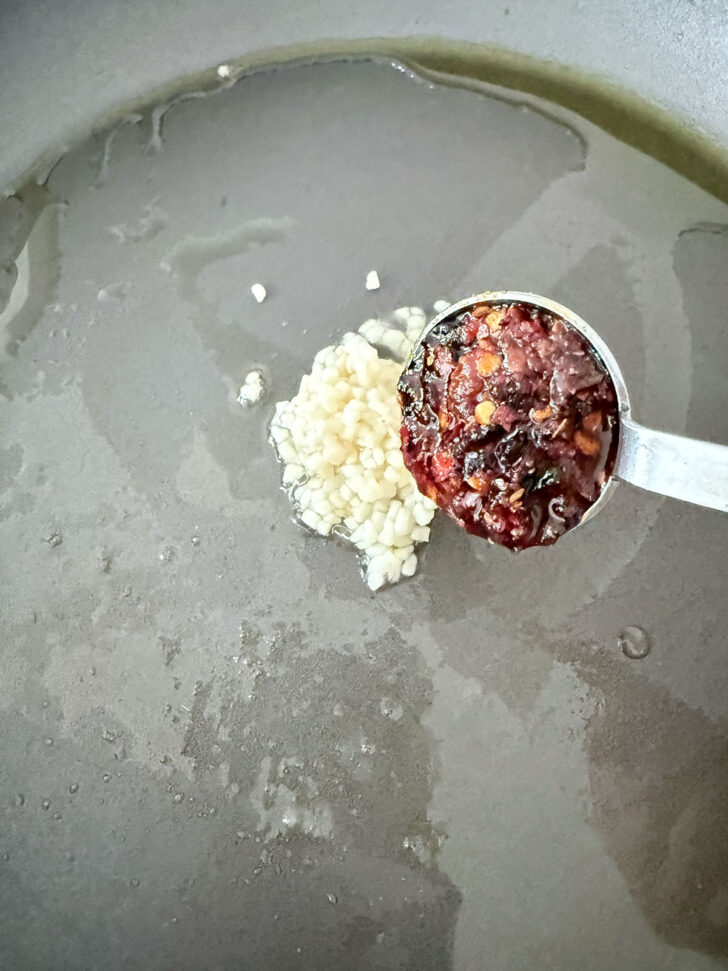 Adding garlic and chili crisp to a hot skillet with oil.
