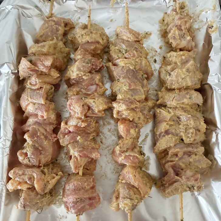Marinated chicken on skewers on a foil-lined baking sheet before baking.