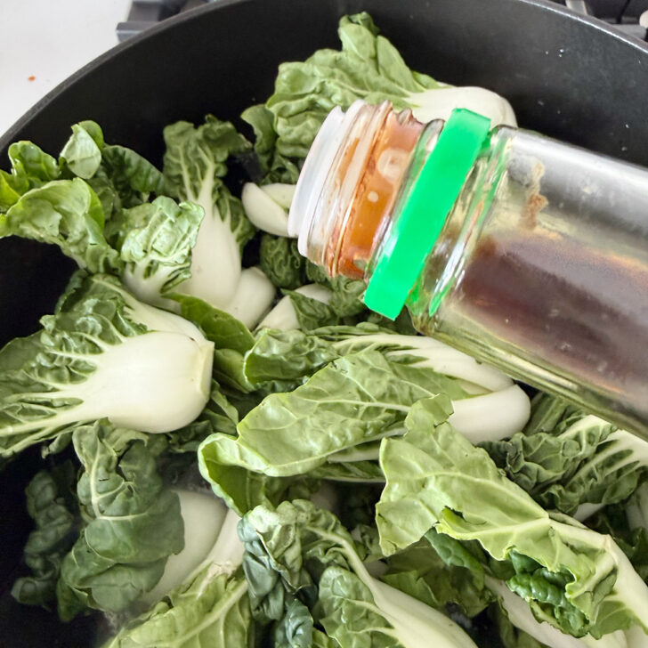 Adding soy sauce to baby bok choy in a skillet.