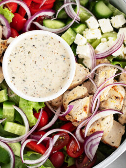 A close-up of a chicken souvlaki salad featuring grilled chicken, cherry tomatoes, cucumber, red onion, lettuce, feta cheese, and a small bowl of creamy dressing in the centre.