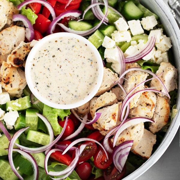 A close-up of a chicken souvlaki salad featuring grilled chicken, cherry tomatoes, cucumber, red onion, lettuce, feta cheese, and a small bowl of creamy dressing in the centre.