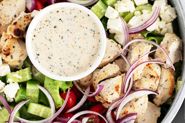 A close-up of a chicken souvlaki salad featuring grilled chicken, cherry tomatoes, cucumber, red onion, lettuce, feta cheese, and a small bowl of creamy dressing in the centre.