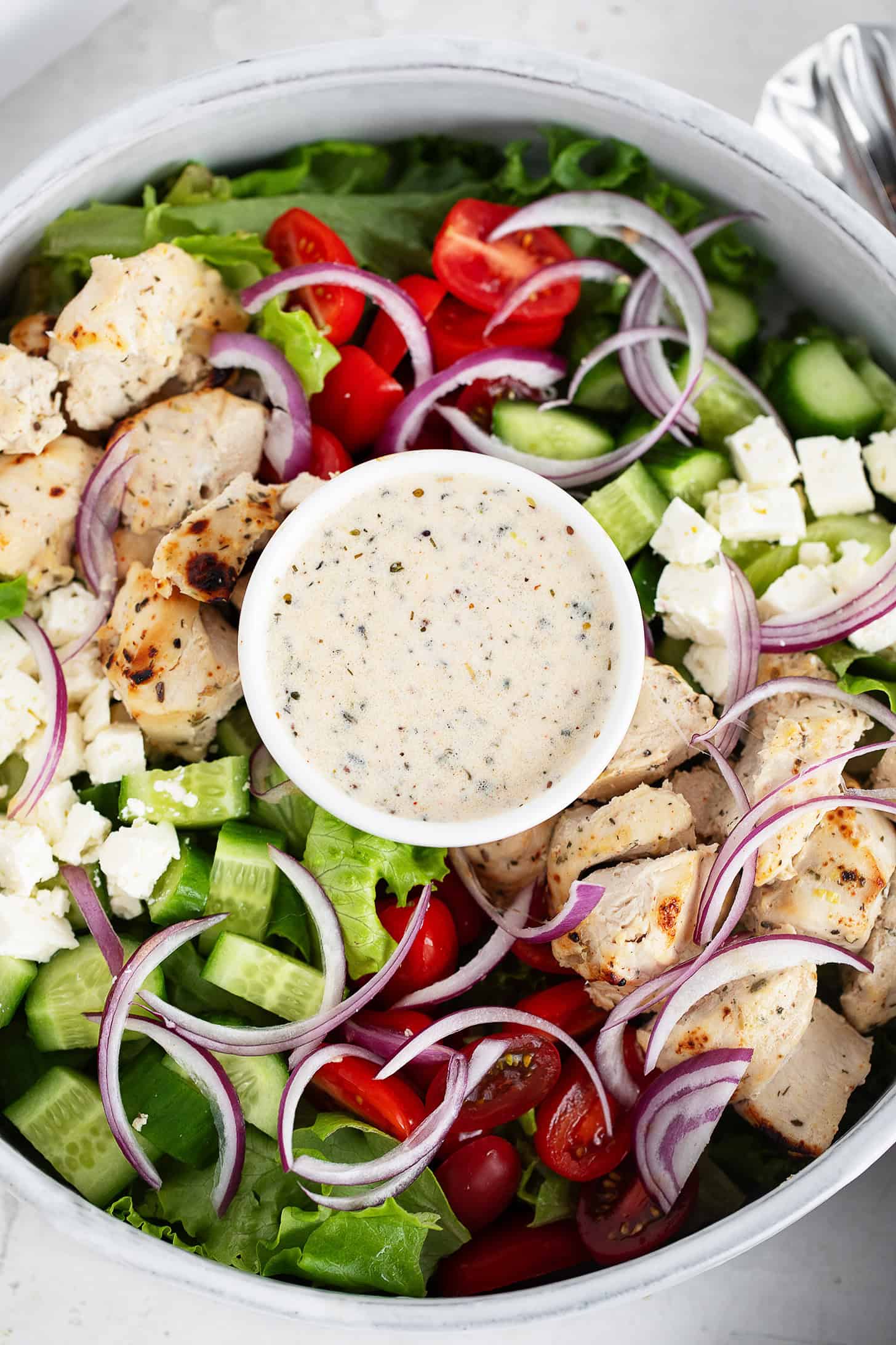 A close-up of a chicken souvlaki salad featuring grilled chicken, cherry tomatoes, cucumber, red onion, lettuce, feta cheese, and a small bowl of creamy dressing in the centre.