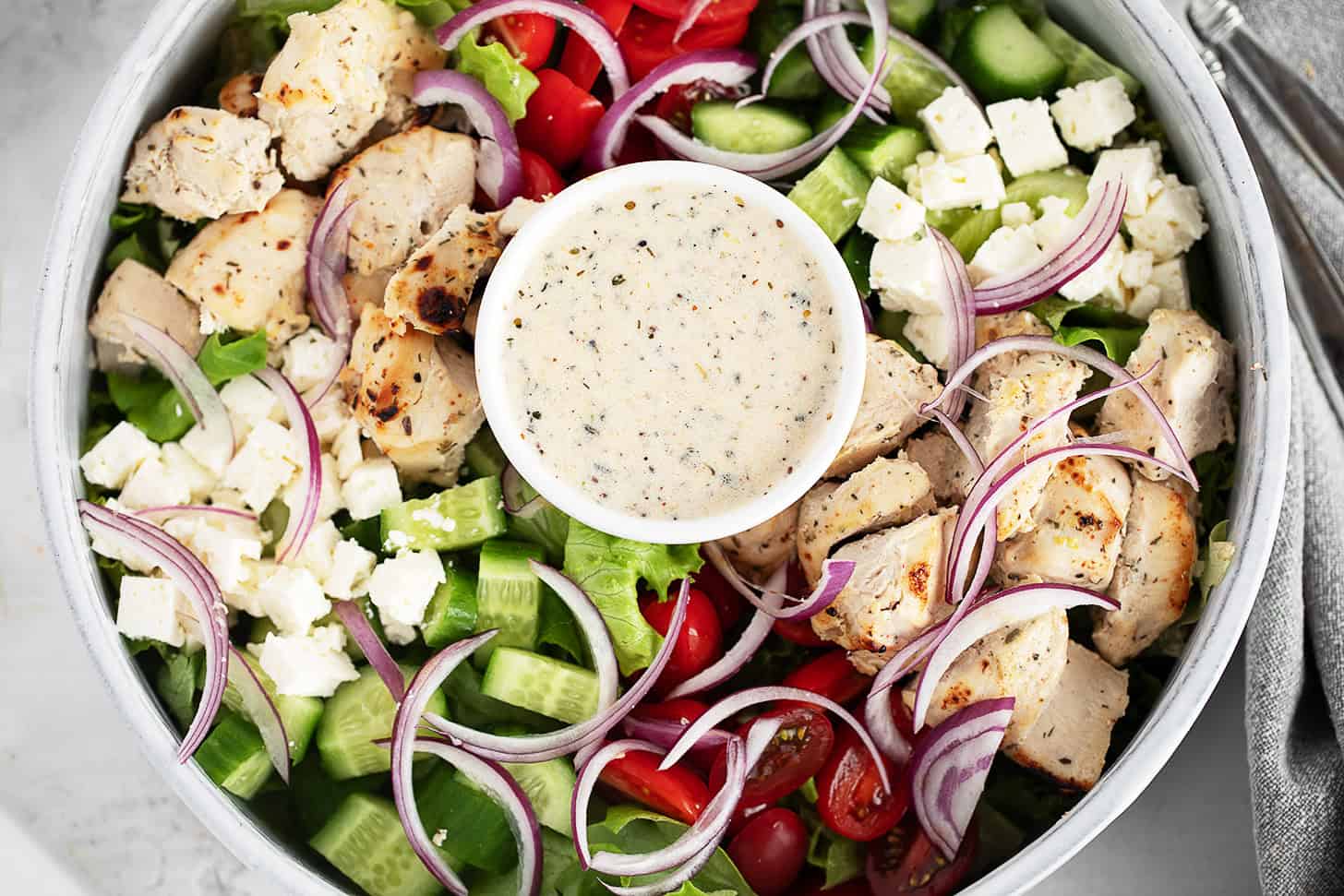 A close-up of a chicken souvlaki salad featuring grilled chicken, cherry tomatoes, cucumber, red onion, lettuce, feta cheese, and a small bowl of creamy dressing in the centre.