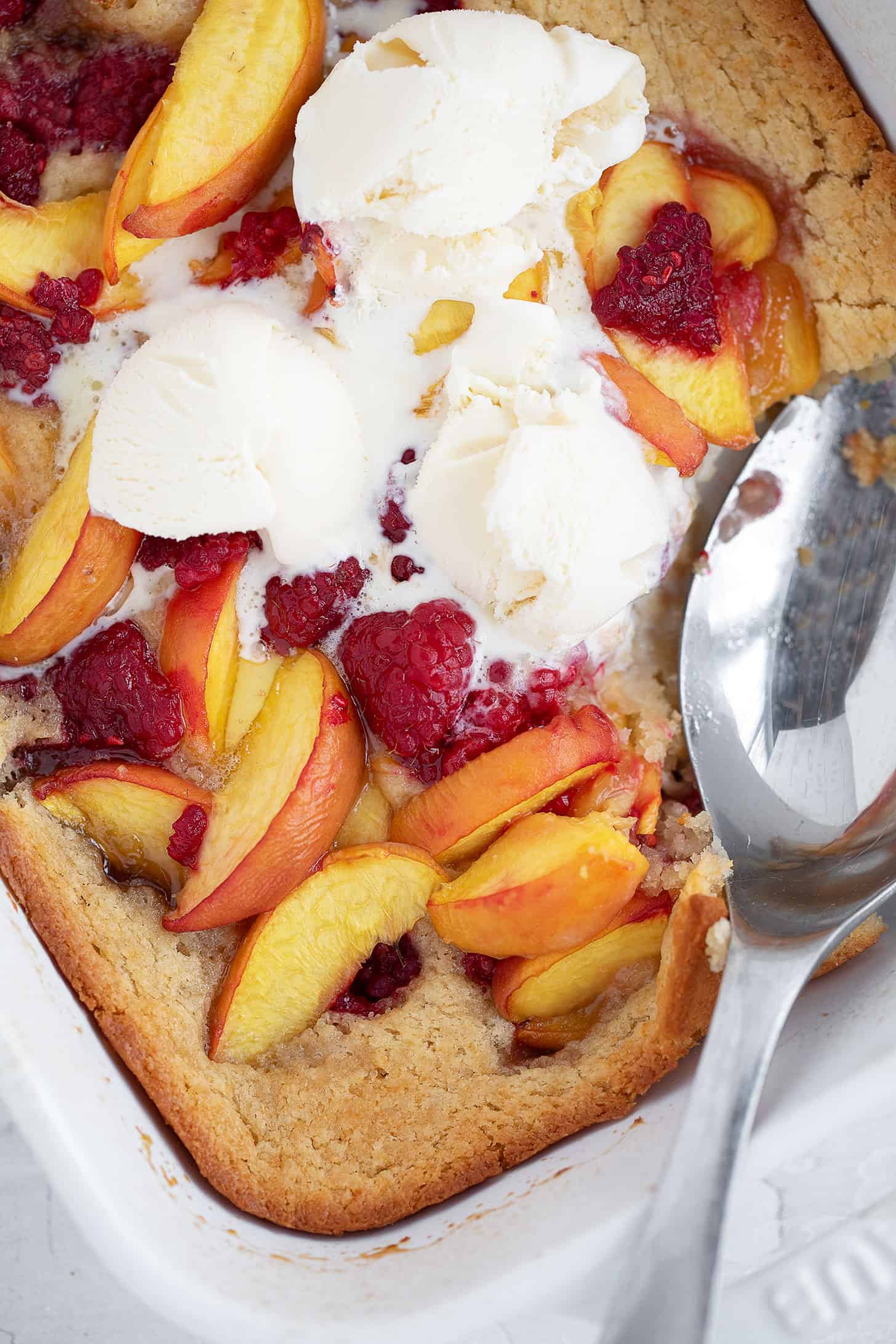Peach melba spoon cake in the baking dish with a spoon and ice cream on top.