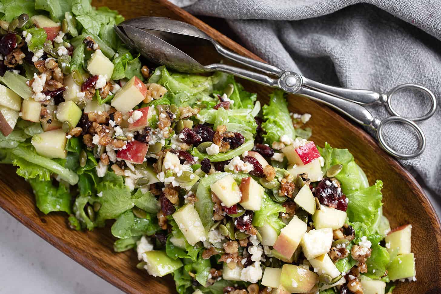 Chopped apple salad in a wooden bowl with silver tongs.