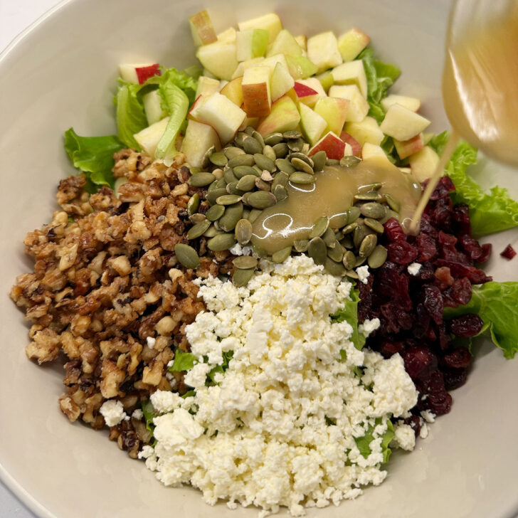 Chopped apple salad ingredients in a large salad bowl with maple mustard vinaigrette being drizzled.