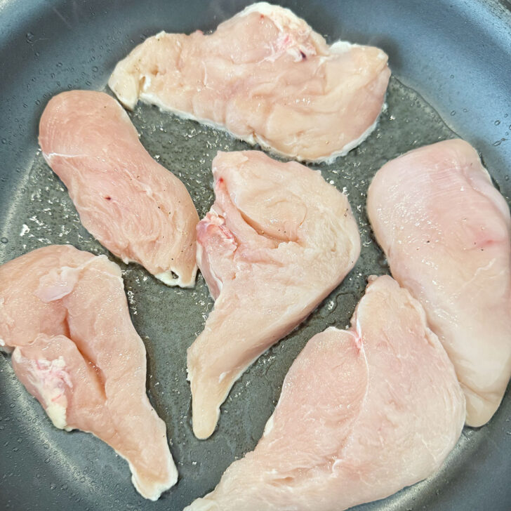 Boneless chicken breast cutlets cooking in a hot skillet with oil.