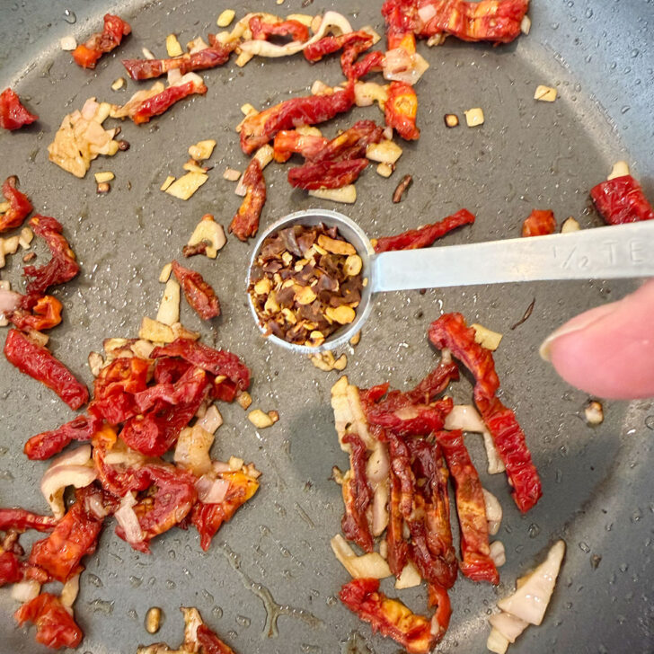 Adding crushed red pepper flakes to a skillet with the shallot and sun-dried tomatoes.